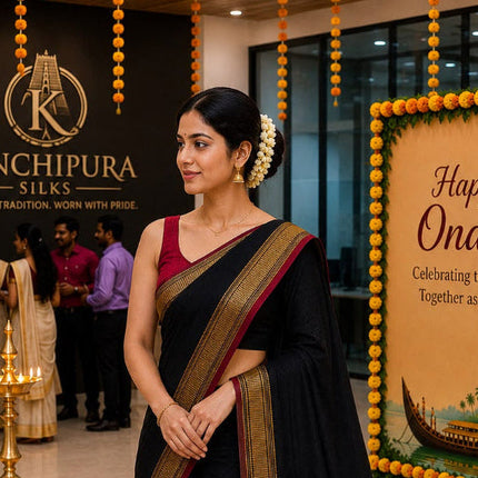 A woman modeling a black silk saree with a deep maroon border and gold zari detailing, standing in a decorated indoor event space with Kanchipura Silks branding and 'Happy Onam' banners.