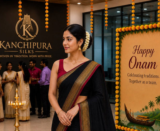 A woman modeling a black silk saree with a deep maroon border and gold zari detailing, standing in a decorated indoor event space with Kanchipura Silks branding and 'Happy Onam' banners.